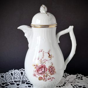 Könnte beinhalten: Weiße Porzellan-Kaffeekanne mit Deckel und Griff. Die Kanne hat ein Blumendesign in Rosa- und Goldtönen. Der Rand der Kanne ist mit Gold verziert. Die Kanne steht auf einer weißen Spitzendecke.