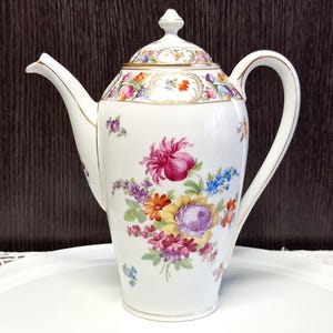 May include: A white porcelain coffee pot with a lid, adorned with colorful floral patterns. The pot features a curved spout, a large handle, and gold trim. The floral design includes pink, purple, orange, and blue flowers. It sits on a white tray.