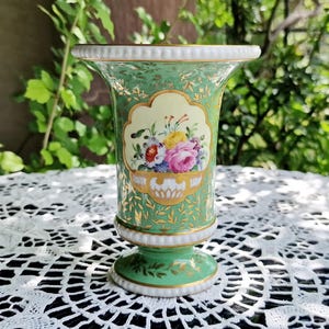 May include: A green and white porcelain vase with a floral design. The vase has a flared top and a pedestal base. A central panel displays a bouquet of colourful flowers. Gold accents adorn the vase.