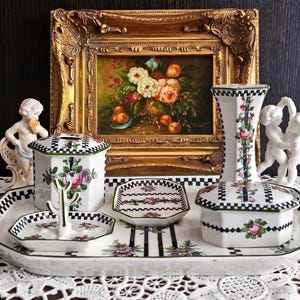 May include: A white ceramic vanity set featuring a black and white checkered border and floral patterns. The set includes an octagonal tray, a lidded container, a ring holder, a small dish, and a tall vase, all decorated with pink roses and green leaves.
