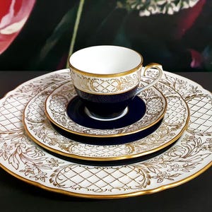 Könnte beinhalten: Ein feines Porzellan-Teeservice mit weißer Tasse, Untertasse und Tellern, verziert mit Goldfiligran und marineblauen Akzenten. Das Set umfasst eine Teetasse, eine Untertasse und zwei Teller.