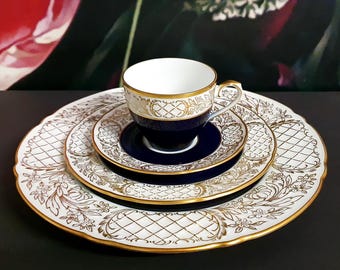 May include: A fine china tea set with a white cup, saucer, and plates, featuring gold filigree and a navy blue accent. The set includes a teacup, a saucer, and two plates, all with a gold trim.