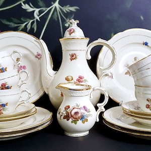 Könnte beinhalten: Ein Vintage-Tee-Set mit cremefarbener Basis und goldener Verzierung. Das Set beinhaltet eine Teekanne, eine Milchkännchen, eine Zuckerdose und sechs Tassen und Untertassen. Die Tassen und Untertassen sind mit einem Blumenmuster mit rosa, roten und blauen Blumen verziert.