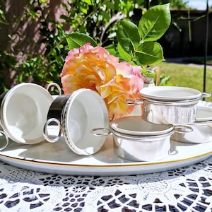 May include: A white and silver tea set on a gold-edged serving tray. The set includes teacups with silver handles and small silver bowls. A bouquet of pink and yellow roses adds colour to the scene. The set is on a white lace tablecloth.