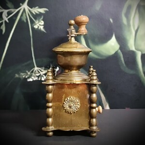May include: Antique brass coffee grinder with a wooden handle and a decorative floral design. The grinder has a square base with ornate details and a small wooden knob on the side.
