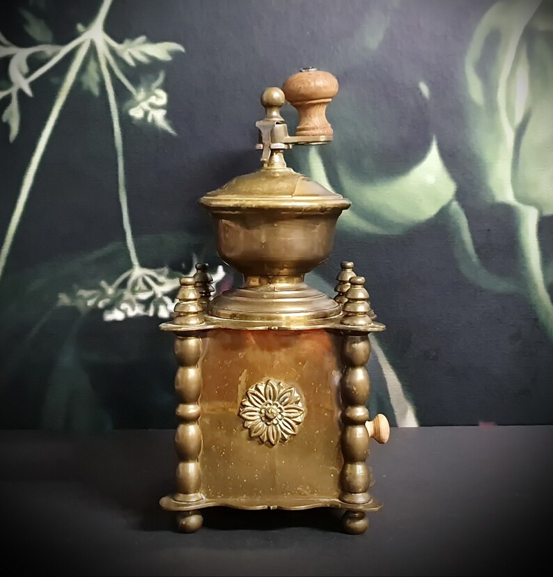 May include: Antique brass coffee grinder with a wooden handle and a decorative floral design. The grinder has a square base with ornate details and a small wooden knob on the side.