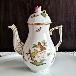 Puede incluir: Tetera de porcelana blanca con ribete dorado, con pájaros, mariposas y motivos florales pintados a mano. La tapa está rematada con una rosa rosa. La tetera tiene un pico curvo y un asa dorada.