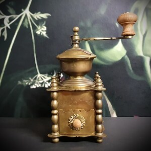 May include: Antique brass coffee grinder with a wooden handle and a decorative floral design. The grinder has a crank and a hopper for coffee beans.