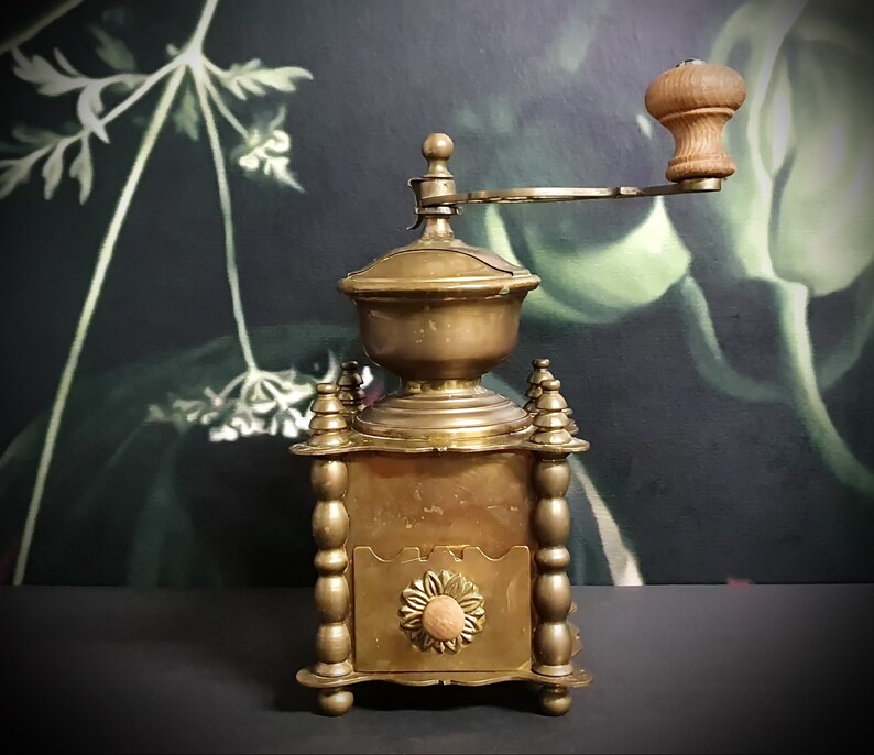 May include: Antique brass coffee grinder with a wooden handle and a decorative floral design. The grinder has a crank and a hopper for coffee beans.