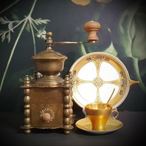 May include: Antique brass coffee grinder with ornate details and a wooden handle. The grinder is on a table with a gold and white porcelain plate and a gold cup.