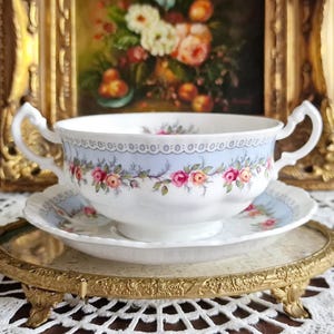 Puede incluir: Un juego de cuenco y platillo de sopa de porcelana blanca con un borde azul claro y un diseño floral. El cuenco tiene dos asas y se apoya en una bandeja dorada. Una pintura enmarcada está en el fondo.