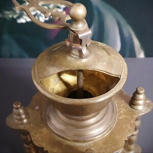 May include: Antique brass coffee grinder with a decorative top and a crank handle. The grinder has a round base with ornate details.