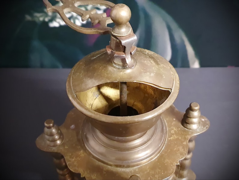May include: Antique brass coffee grinder with a decorative top and a crank handle. The grinder has a round base with ornate details.