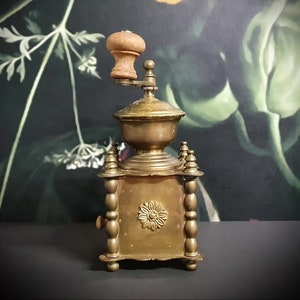 May include: Antique brass coffee grinder with a wooden handle and intricate details. The grinder has a square base with four decorative columns and a round top with a floral design.