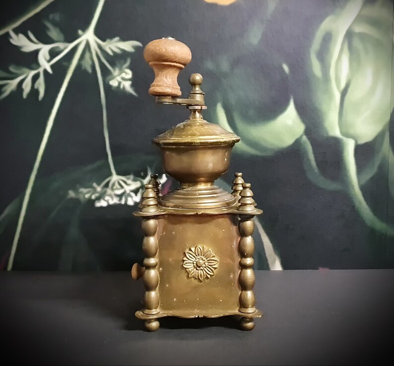 May include: Antique brass coffee grinder with a wooden handle and intricate details. The grinder has a square base with four decorative columns and a round top with a floral design.
