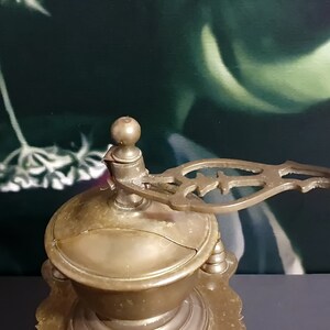 May include: Antique brass coffee grinder with a wooden handle and intricate details. The grinder has a round, domed top and a base with a decorative design.