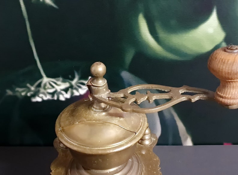 May include: Antique brass coffee grinder with a wooden handle and intricate details. The grinder has a round, domed top and a base with a decorative design.