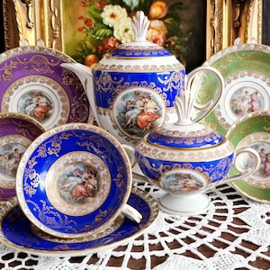 Könnte beinhalten: Ein Teeservice aus Porzellan bestehend aus einer Teekanne, einer Zuckerdose mit Deckel sowie vier Untertassen mit passenden Tassen. Die Stücke sind in leuchtendem Blau, Violett und Grün mit goldenen Filigranverzierungen und zentralen Medaillons.