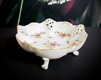 Puede incluir: Un tazón de porcelana blanca con un borde dorado y un patrón floral. El tazón tiene tres patas y está decorado con pequeñas rosas rosas. Tiene un borde festoneado y un diseño calado.