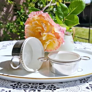 May include: A white and gold serving tray holds two silver teacups and a small creamer. A large, multi-coloured rose and greenery are in a small vase. The scene is set on a white lace tablecloth.