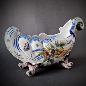 May include: A white ceramic planter with a scalloped rim and three feet. The planter is decorated with blue, yellow, and red floral and butterfly designs. The ends of the planter have a scrolled design.