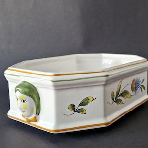 Może przedstawiać: Biała prostokątna ceramiczna donica z malowanym kwiatowym wzorem i twarzą kobiety z boku. Donica ma żółto-zielone obramowanie i ma około 25 cm długości.
