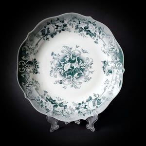 Puede incluir: Un plato de cerámica blanco con un diseño floral verde. El plato tiene un borde festoneado y está decorado con un motivo floral central y un borde de hojas y flores.
