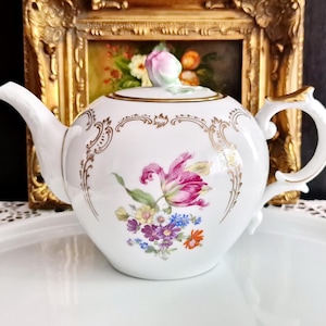 Può includere: Una teiera in porcellana bianca con un motivo floreale dipinto a mano che include un tulipano rosa e piccoli fiori colorati. La teiera è decorata con volute dorate, un manico con bordo in oro e un coperchio sormontato da un bocciolo di rosa scolpito.