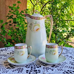 May include: A porcelain coffee set comprising a tall pitcher and two matching cups with saucers. The items feature a pale green base, hand-painted yellow and pink rose floral motifs, and gold-trimmed rims and handles on the pitcher and cups.