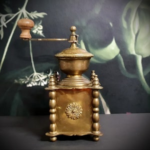 May include: Antique brass coffee grinder with a wooden handle and ornate details. The grinder has a square base with decorative columns and a round top.