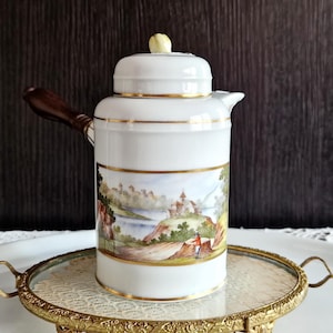 Può includere: Caffettiera in ceramica bianca con scena di paesaggio, bordi dorati e manico in legno. La caffettiera ha un coperchio con un piccolo pomello giallo. È appoggiata su un vassoio con bordo dorato e piano in vetro. La scena raffigura un castello e figure.