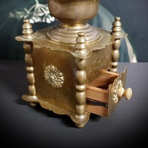 May include: Antique brass coffee grinder with a wooden drawer and a decorative floral design. The grinder has a square base and a round top.
