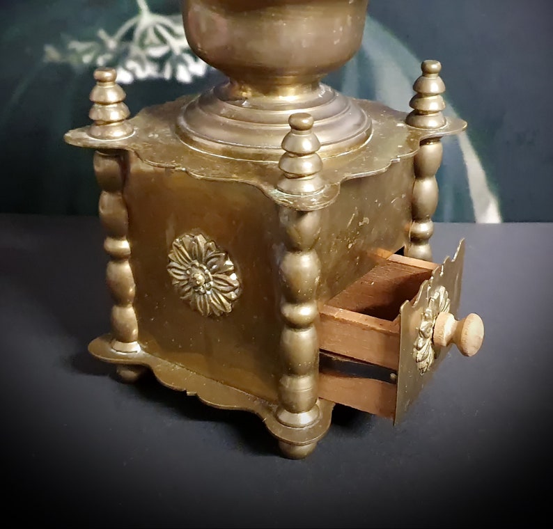 May include: Antique brass coffee grinder with a wooden drawer and a decorative floral design. The grinder has a square base and a round top.