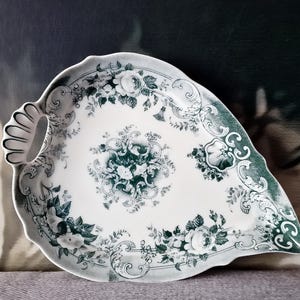 Puede incluir: Un plato de servir de porcelana blanca con un diseño floral verde. El plato tiene forma de hoja y un asa en el lateral.