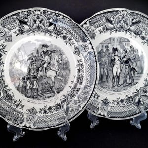 May include: Two black and white porcelain plates with decorative borders. One plate features a scene of a military leader on horseback, while the other depicts a group of soldiers in uniform.