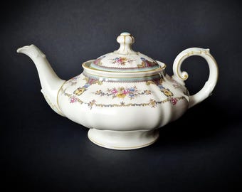 May include: Cream-colored porcelain teapot with a floral design. The teapot features a curved spout, a rounded body, and a lid with a decorative knob. Gold trim accents the handle, spout, and lid. The floral pattern includes pink, blue, and yellow flowers.