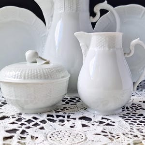 Könnte beinhalten: Ein weißes Porzellan-Teeservice, bestehend aus einer Zuckerdose mit Deckel, einem Sahnekännchen, einer Kanne und zwei Tellern. Das Set hat ein strukturiertes Muster und ist auf einer weißen Spitzendecke arrangiert. Der Hintergrund ist schwarz.