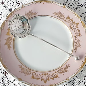 May include: A silver tea infuser with a decorative, perforated bowl and a twisted handle rests on a pink and white china plate. The plate has a gold floral design around the rim.