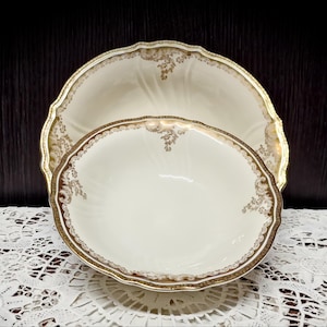 Può includere: Servizio da portata antico in porcellana in due pezzi. Il set comprende un grande piatto con bordo smerlato e una ciotola ovale più piccola. Entrambi i pezzi sono color avorio con bordi dorati e decorazioni floreali. Presentato su una tovaglia di pizzo bianco.