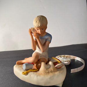 May include: A porcelain figurine of a young boy sitting on a beach, holding a seashell to his ear. He is wearing a blue and white swimsuit and is surrounded by sand, a bucket, a shovel, and a seashell.