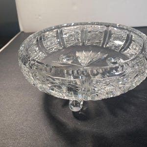 May include: A clear glass bowl with a cut glass design. The bowl has three legs and is sitting on a dark surface.