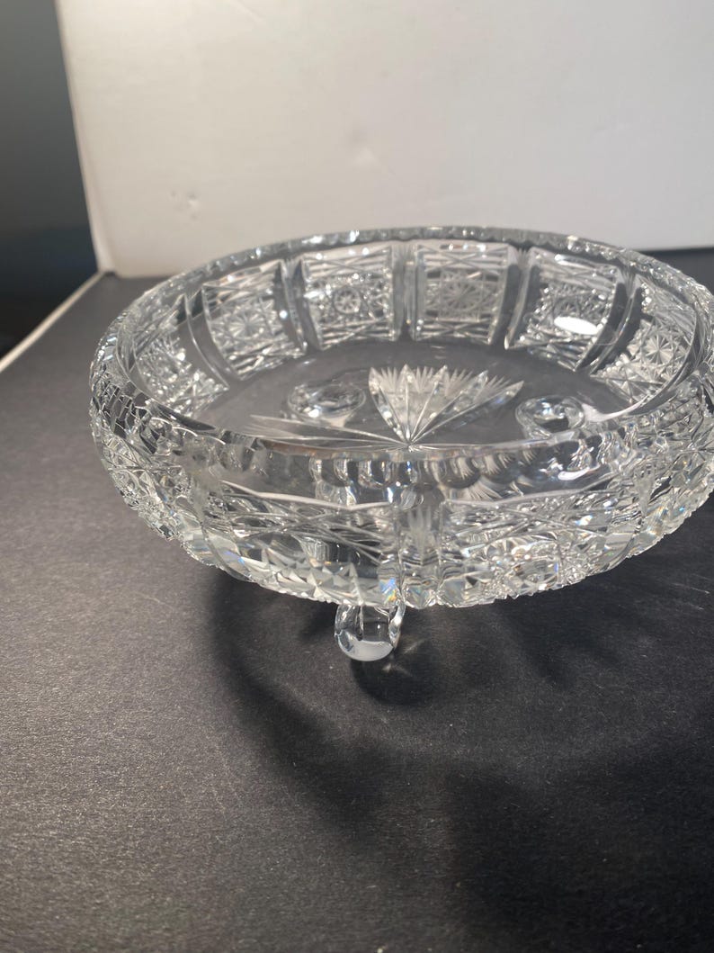 May include: A clear glass bowl with a cut glass design. The bowl has three legs and is sitting on a dark surface.