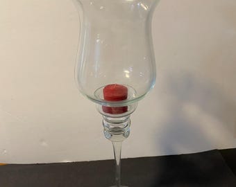 May include: A tall, clear glass candle holder with a wide, flared top and a single red candle inside. The holder has a round base and a long, slender stem.