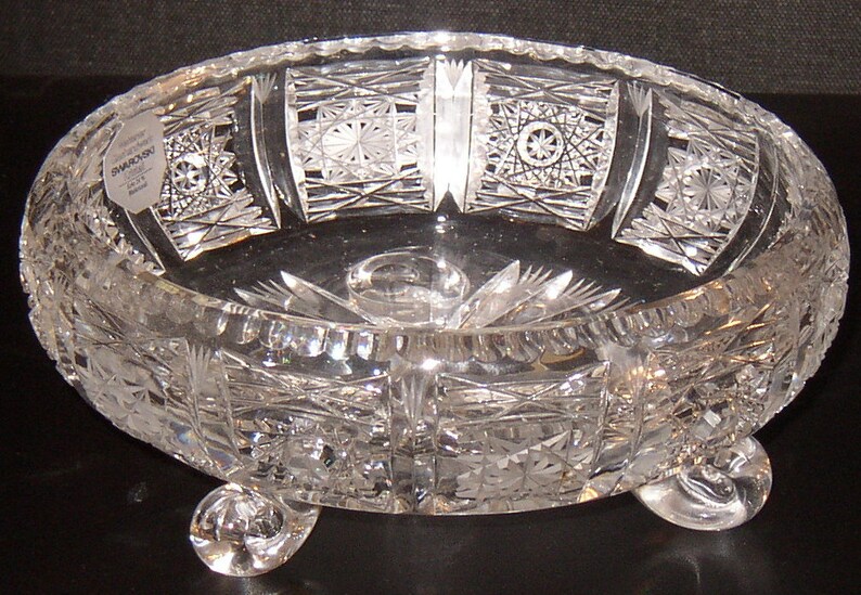 May include: A clear glass bowl with intricate cut designs and three feet. The bowl has a label that reads "Swarovski" and "Made in Austria".