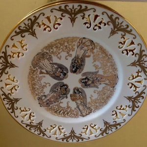 Op de afbeelding: Een wit porseleinen bord met een gouden rand en ingewikkelde gouden bloemmotieven. In het midden bevindt zich een cirkelvormig ontwerp met vijf engelen in goud en wit, omringd door een wervelend patroon. Het jaar 2000 is onderaan in goud gedrukt.
