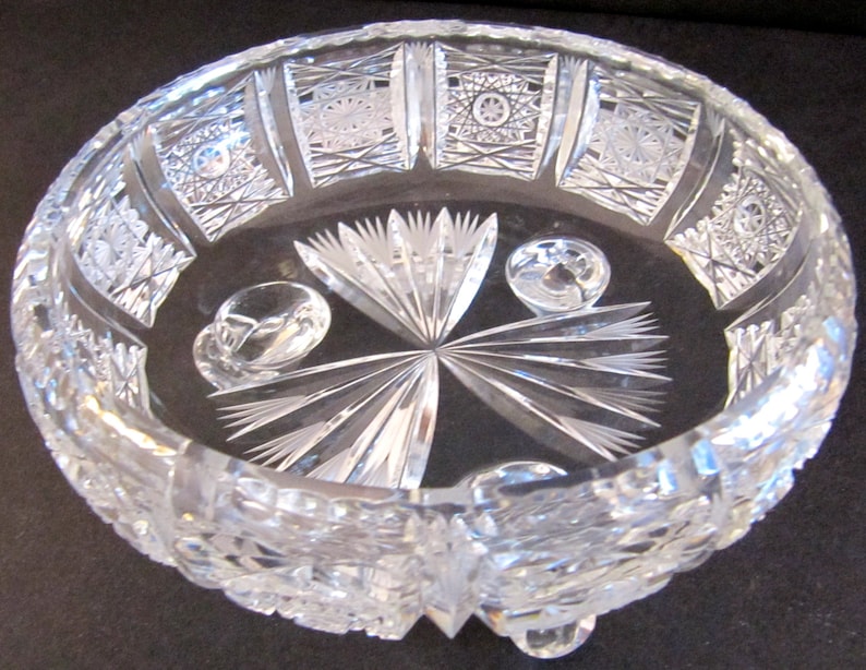 May include: A clear glass bowl with intricate cut designs and three feet. The bowl has a starburst pattern in the centre and a geometric pattern around the rim.