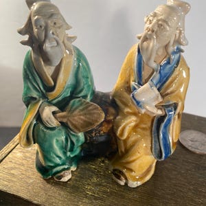 May include: A ceramic figurine depicting two elderly Asian figures seated together. One figure wears a green robe and holds a fan, while the other is in a yellow robe with blue accents. The figures have long white beards and are seated on a dark base.