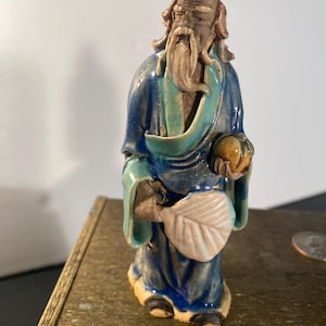 Op de afbeelding: Een klein keramiek beeldje van een oude man met een lange witte baard, die een blauwe en groene robe draagt, een perzik en een waaier vasthoudt.
