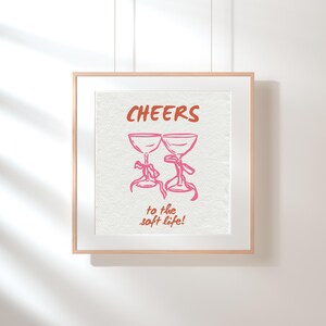 Digital wall art with pink illustrated champagne glasses and red text that says “Cheers to the soft life!” on a textured cream background.