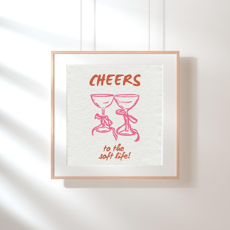 Digital wall art with pink illustrated champagne glasses and red text that says “Cheers to the soft life!” on a textured cream background.